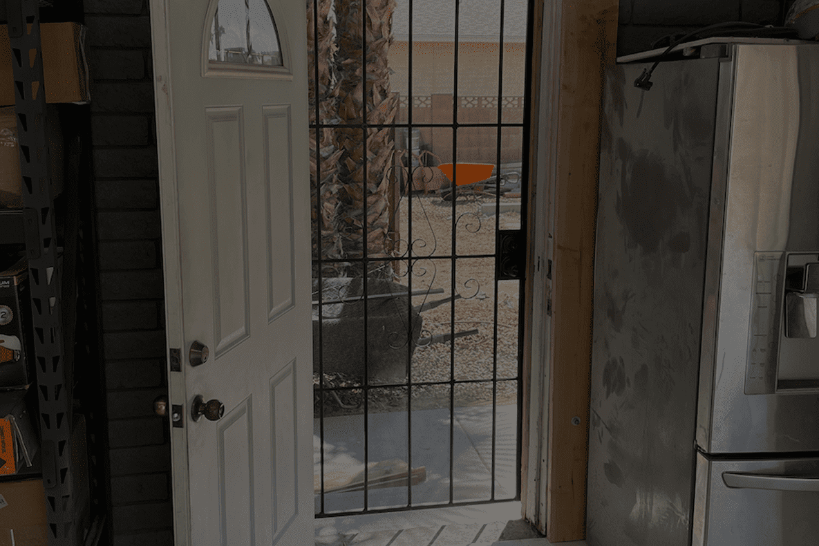 A white door with a black security screen door opens to a sunlit outdoor area with a trash bin.